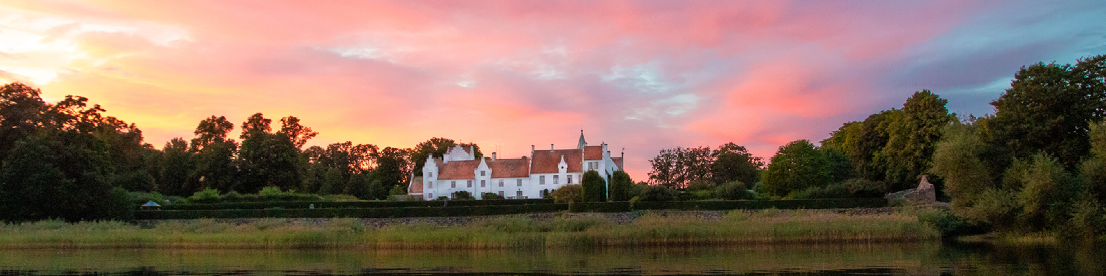 Hvidt slot ved søbred i solnedgang med farverige skyer og omgivet af grønne træer og buske.