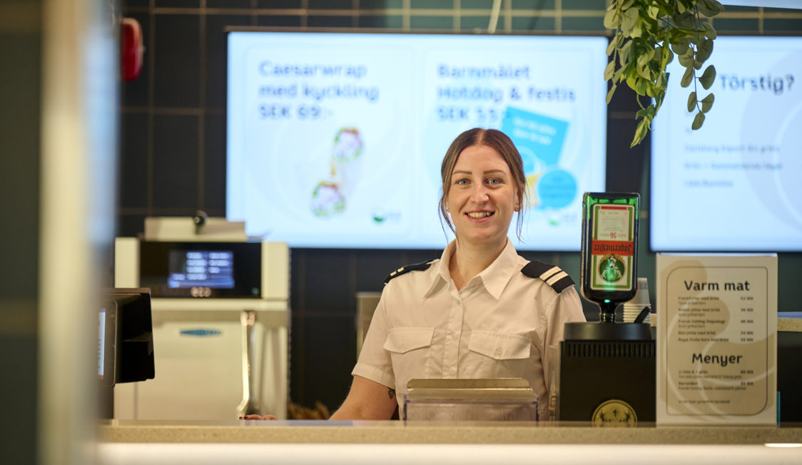 Kvinna i uniform står leende vid kassan i ett café med menytavla och kaffemaskin i bakgrunden.