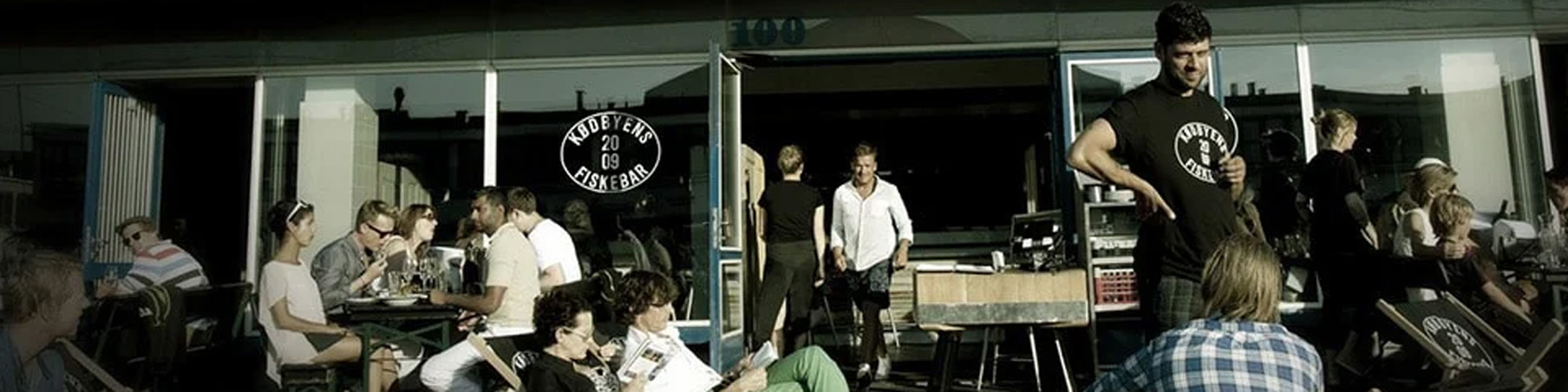 Människor njuter av soligt väder utanför en restaurang i Köpenhamn, Kodbyen Fiskehal i bakgrunden.