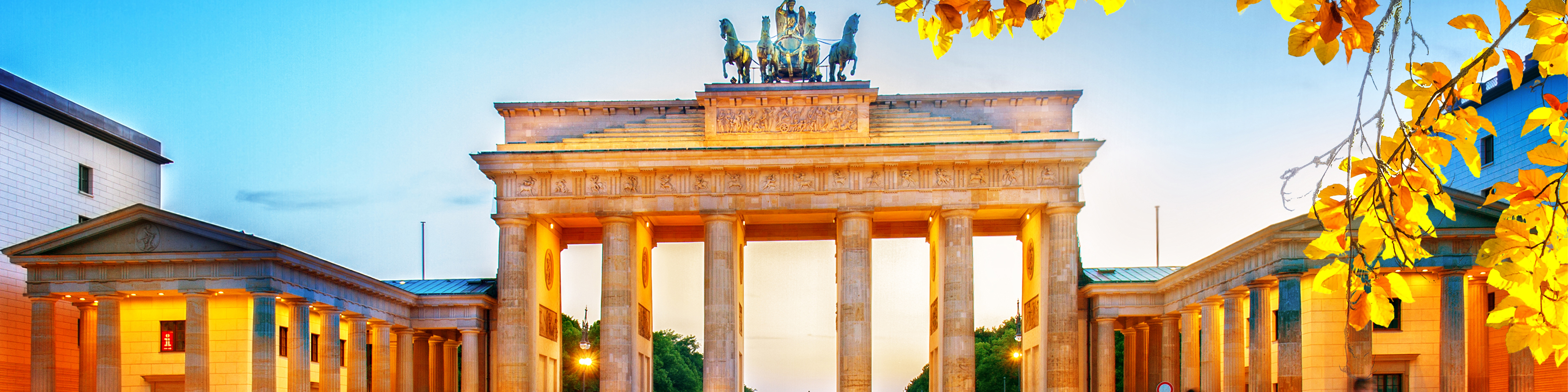 Brandenburger Tor upplyst i skymningen med gyllene löv i förgrunden och klar himmel i bakgrunden.