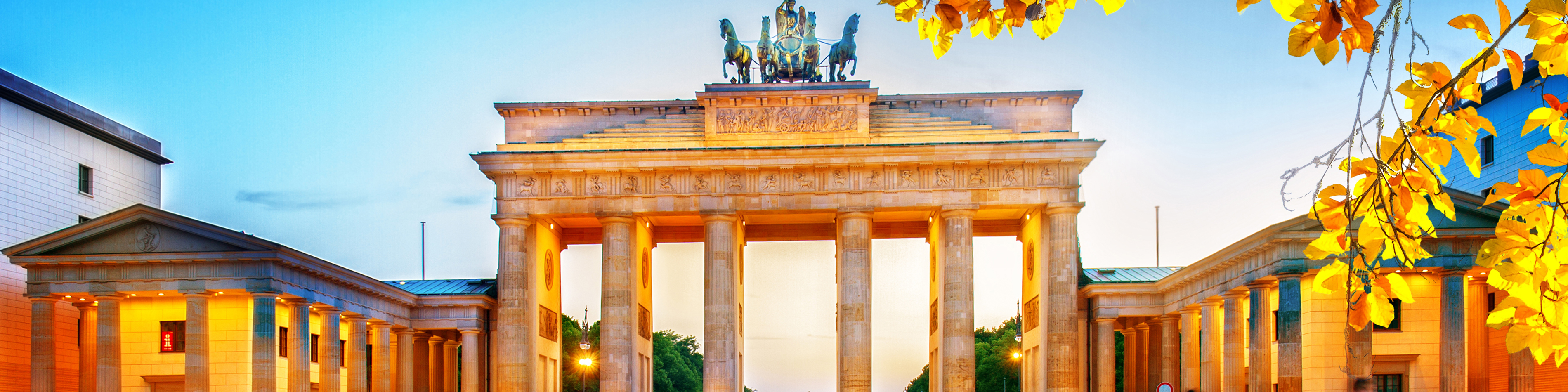 Brandenburger Tor upplyst i skymningen med gyllene löv i förgrunden och klar himmel i bakgrunden.