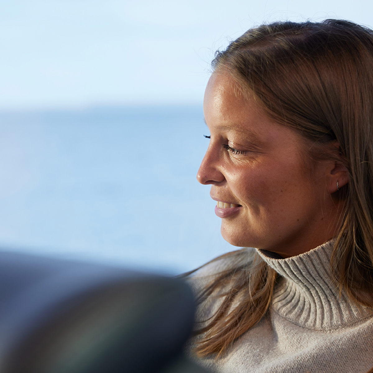 Kvinna i beige stickad tröja tittar ut mot havet, belysning från sidan ger ett mjukt intryck.