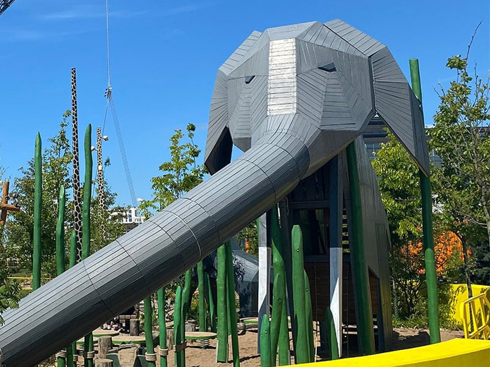 Elefantformet rutchebane i træ og metal i en legeplads omgivet af grønne træskulpturer og planter.