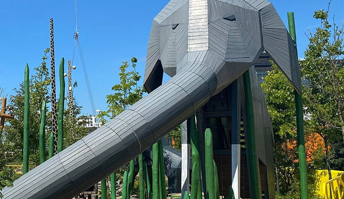 Elefantformet rutchebane i træ og metal i en legeplads omgivet af grønne træskulpturer og planter.