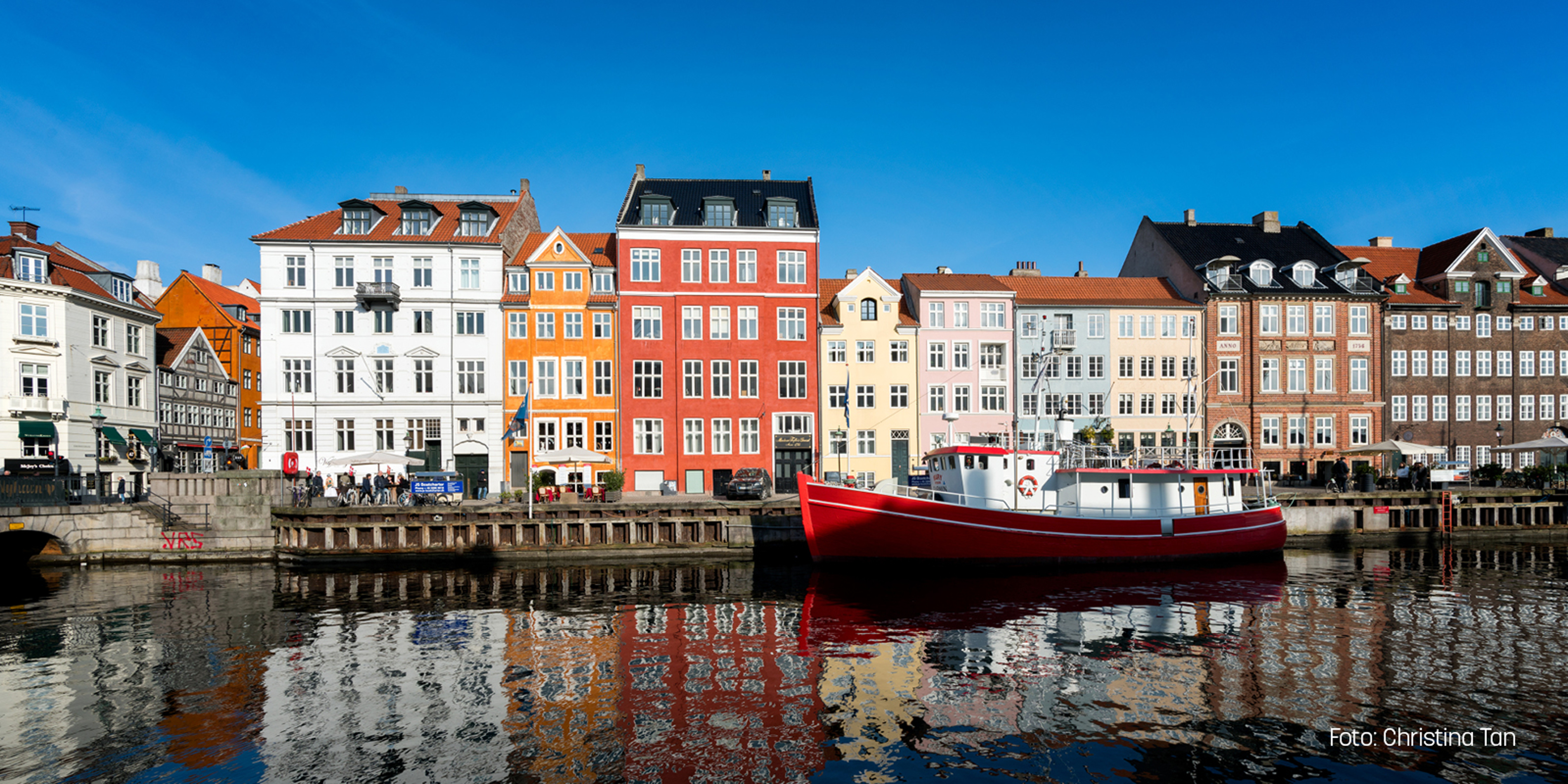 Färgglada husfasader vid kanalen i Köpenhamn med röd båt i förgrunden under klarblå himmel.