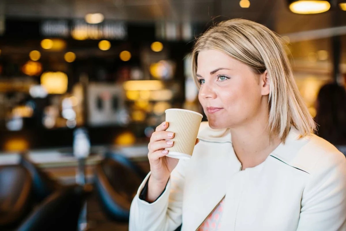 Kvinde i hvid jakke drikker kaffe fra papbæger ved cafébord.