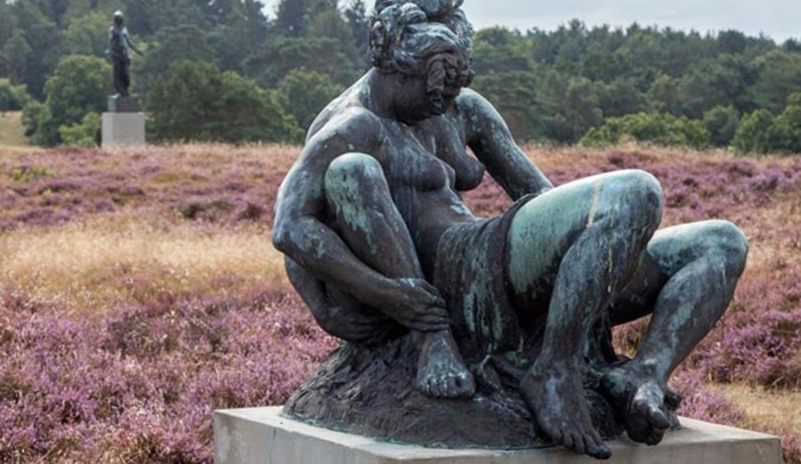 Staty av Rudolph Tegner i brons på Dronningmølle omgiven av lila ljung och skog i bakgrunden.