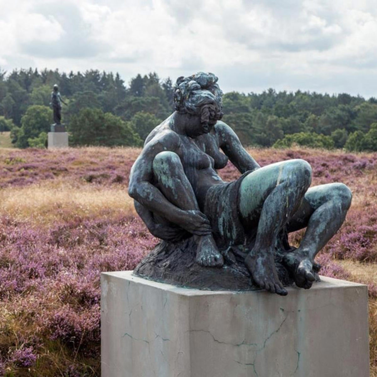 Staty av Rudolph Tegner i brons på Dronningmølle omgiven av lila ljung och skog i bakgrunden.