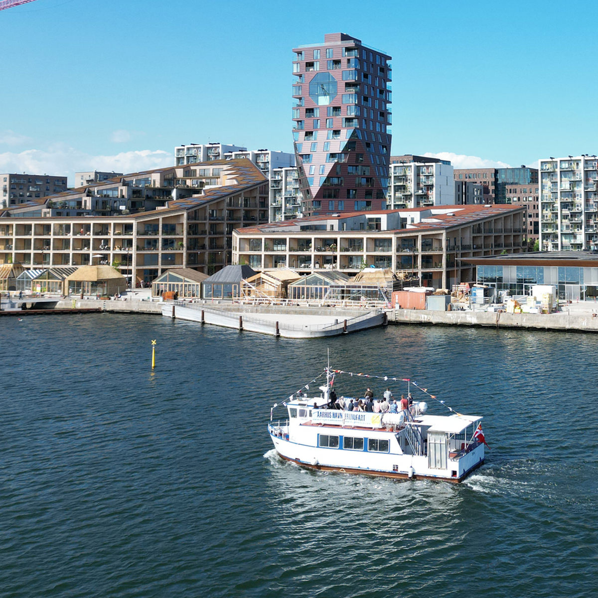 Bybåde Aarhus sejler forbi moderne lejlighedskompleks med unik arkitektur ved havnen under en klar blå himmel.