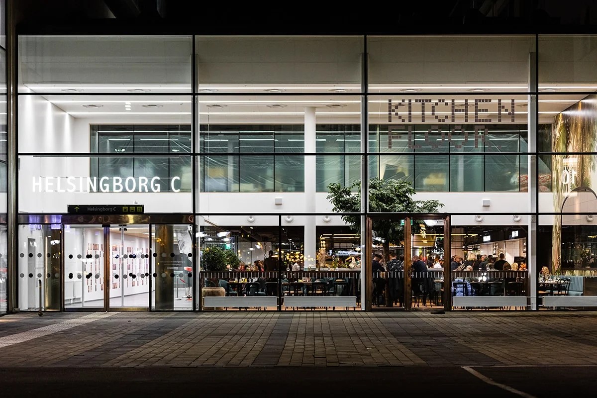 Helsingborg C indgang set udefra om aftenen med folk, der spiser i restauranten Kitchen and Table bag glasfacade.