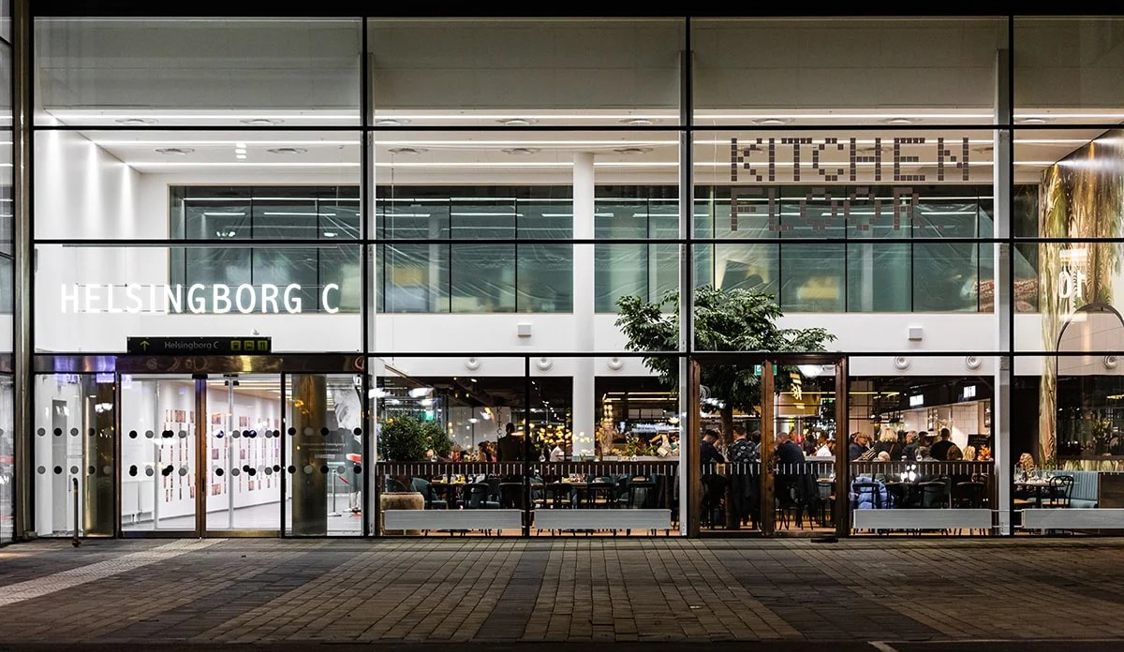 Helsingborg C indgang set udefra om aftenen med folk, der spiser i restauranten Kitchen and Table bag glasfacade.