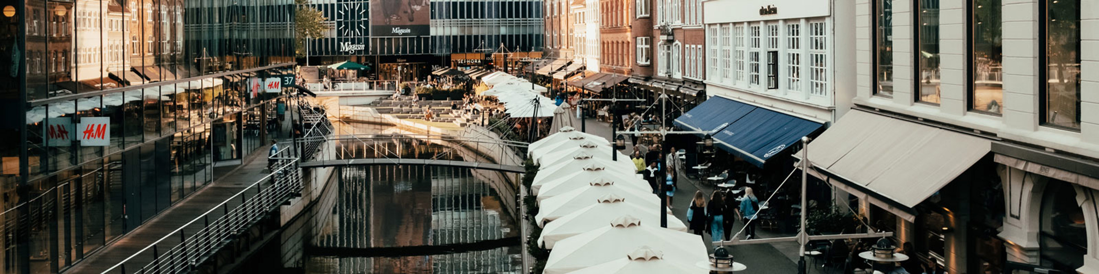 Butiksgade med farverige bygninger og travle caféer langs kanalen i Aarhus set fra oven.