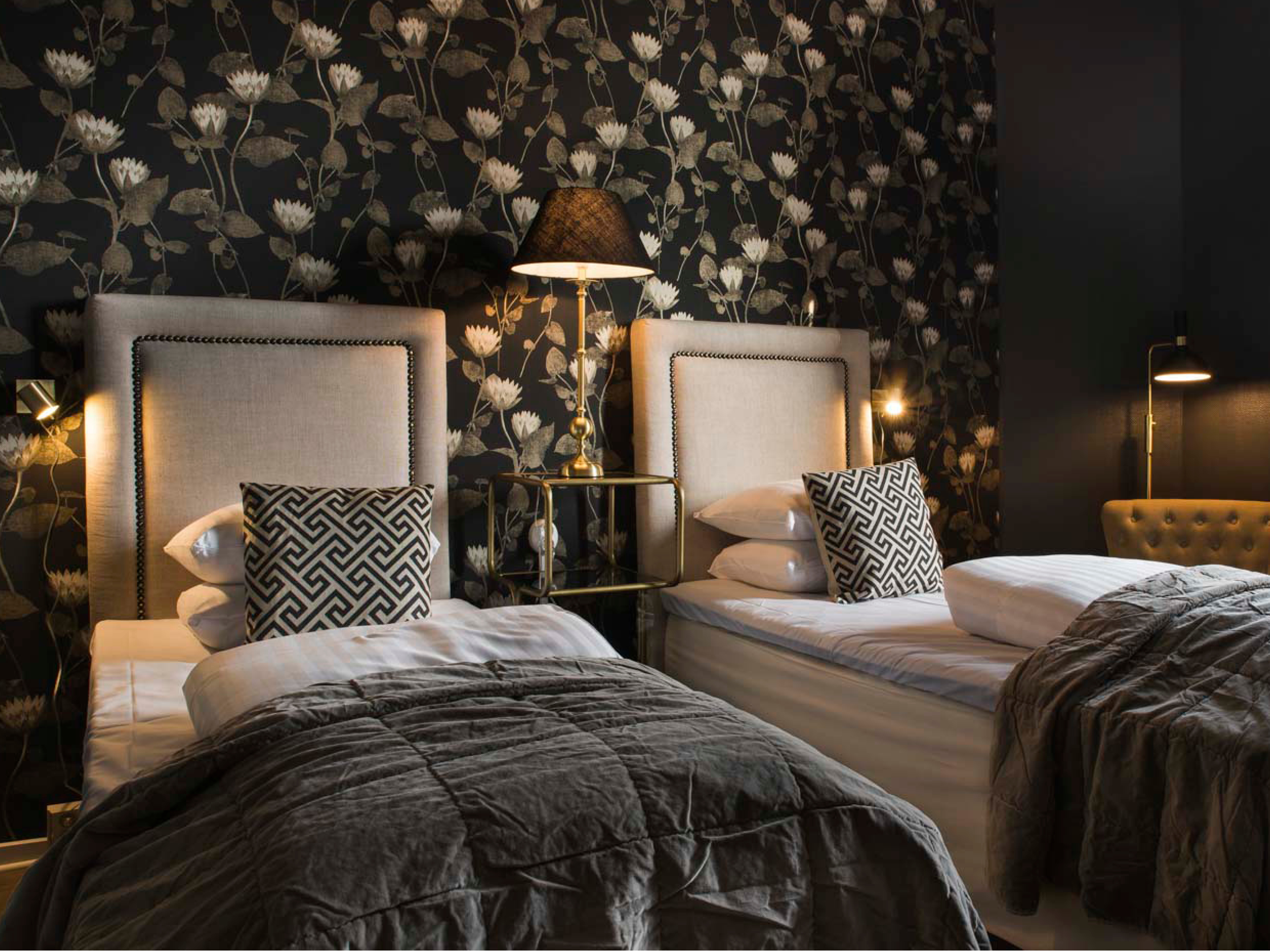 To enkeltsenge med beige sengegavl i værelse med sort blomstret tapet og elegante puder på hver seng.