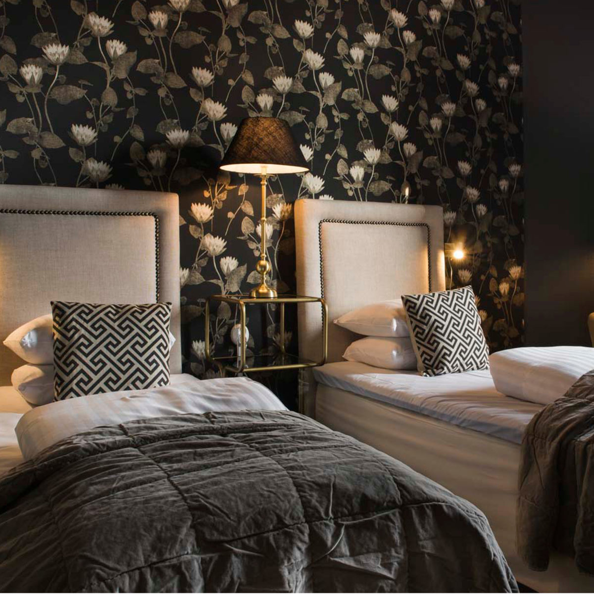 To enkeltsenge med beige sengegavl i værelse med sort blomstret tapet og elegante puder på hver seng.