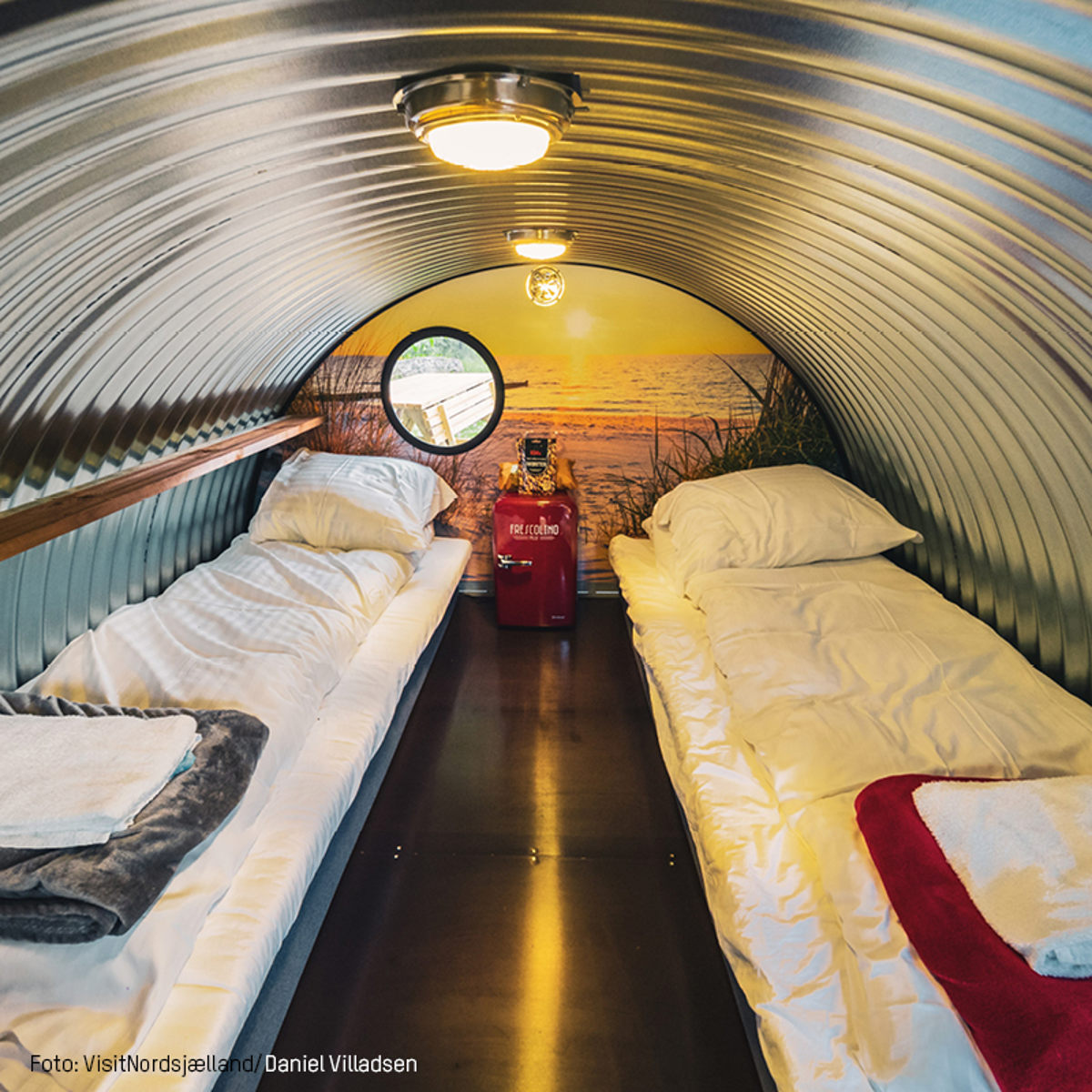 Interiør af et rørhotel med to enkeltsenge, rund ventilation i loftet og vægmaleri af solopgang ved havet.