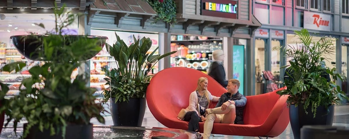 To mennesker sidder på en rød moderne sofa i et indkøbscenter, omgivet af grønne planter i krukker.