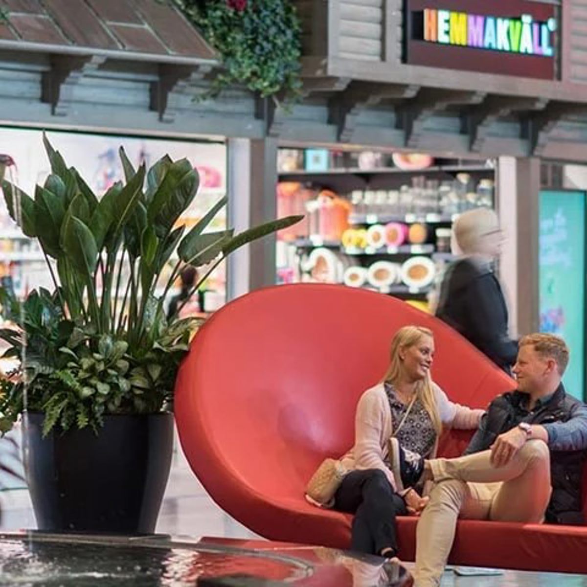 To mennesker sidder på en rød moderne sofa i et indkøbscenter, omgivet af grønne planter i krukker.