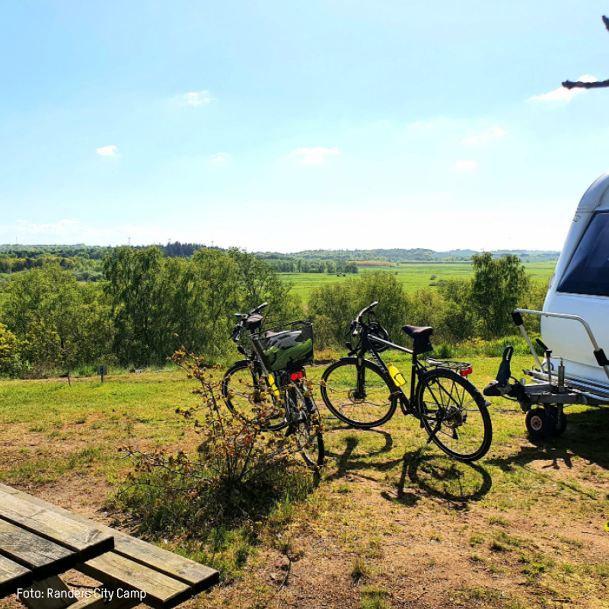To cykler parkeret ved campingvogn i frodigt landskab ved Randers City Camp på solrig dag.