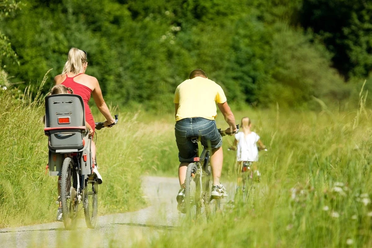 Familjecykling på landsväg med barnstol, sommarklädda föräldrar och barn i grönt landskap.