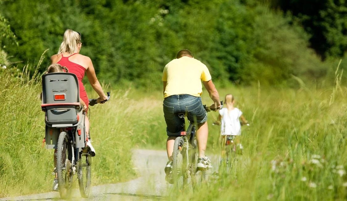 Familjecykling på landsväg med barnstol, sommarklädda föräldrar och barn i grönt landskap.