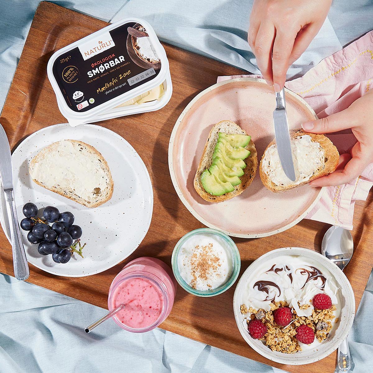Økologisk smørbar og skiver af brød med avocado og smørepålæg på træbakke ved siden af skål med yoghurt og bær.
