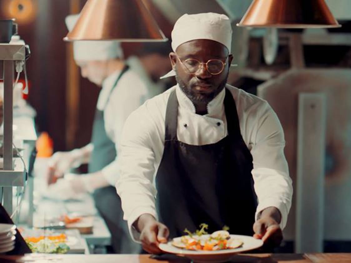 Kock i vit uniform och svart förkläde serverar en färgglad maträtt på en restaurang i köket.