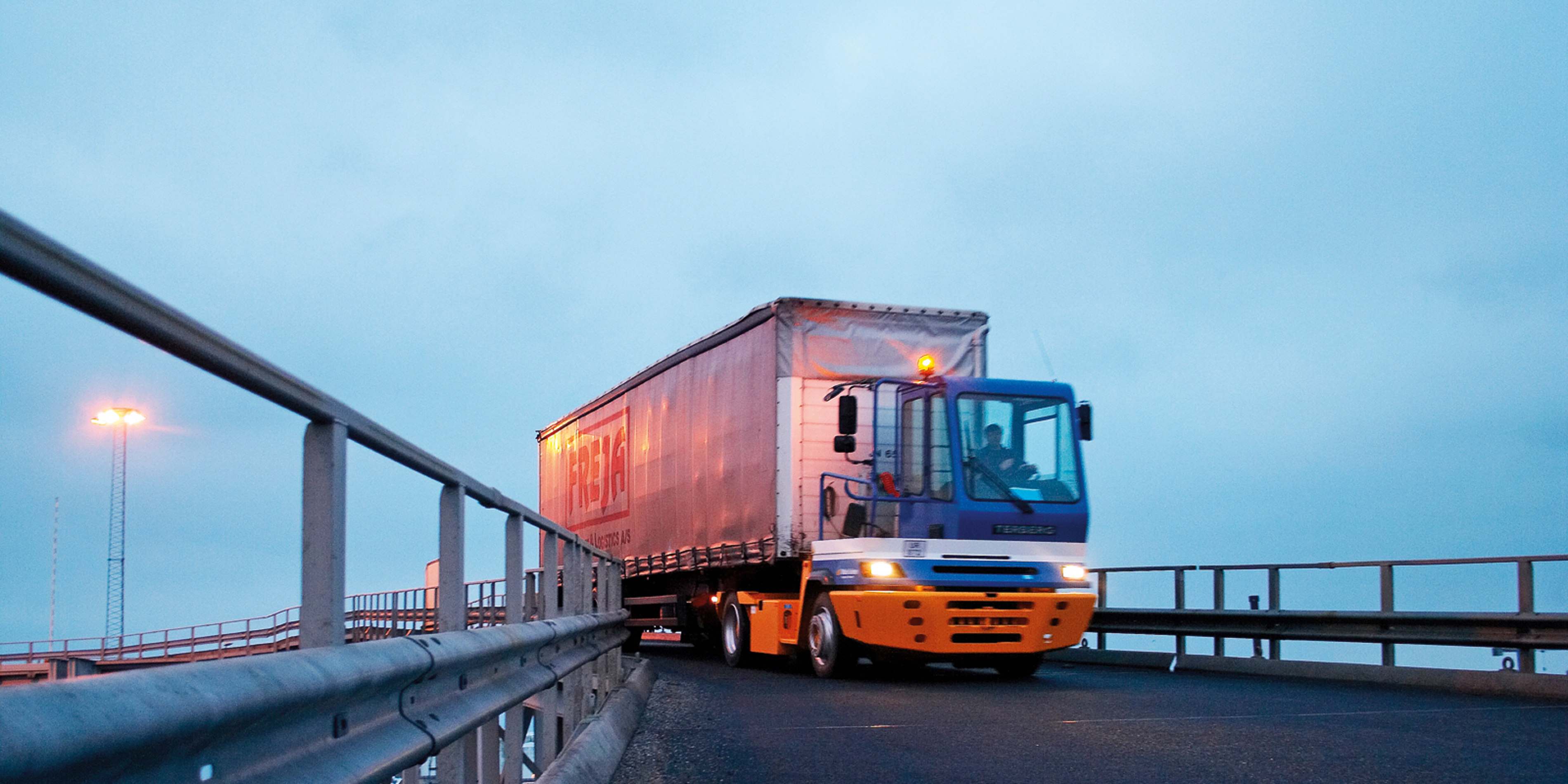 Fraktbil med container kör upp på färjan under klarblå himmel, sedd från sidan.