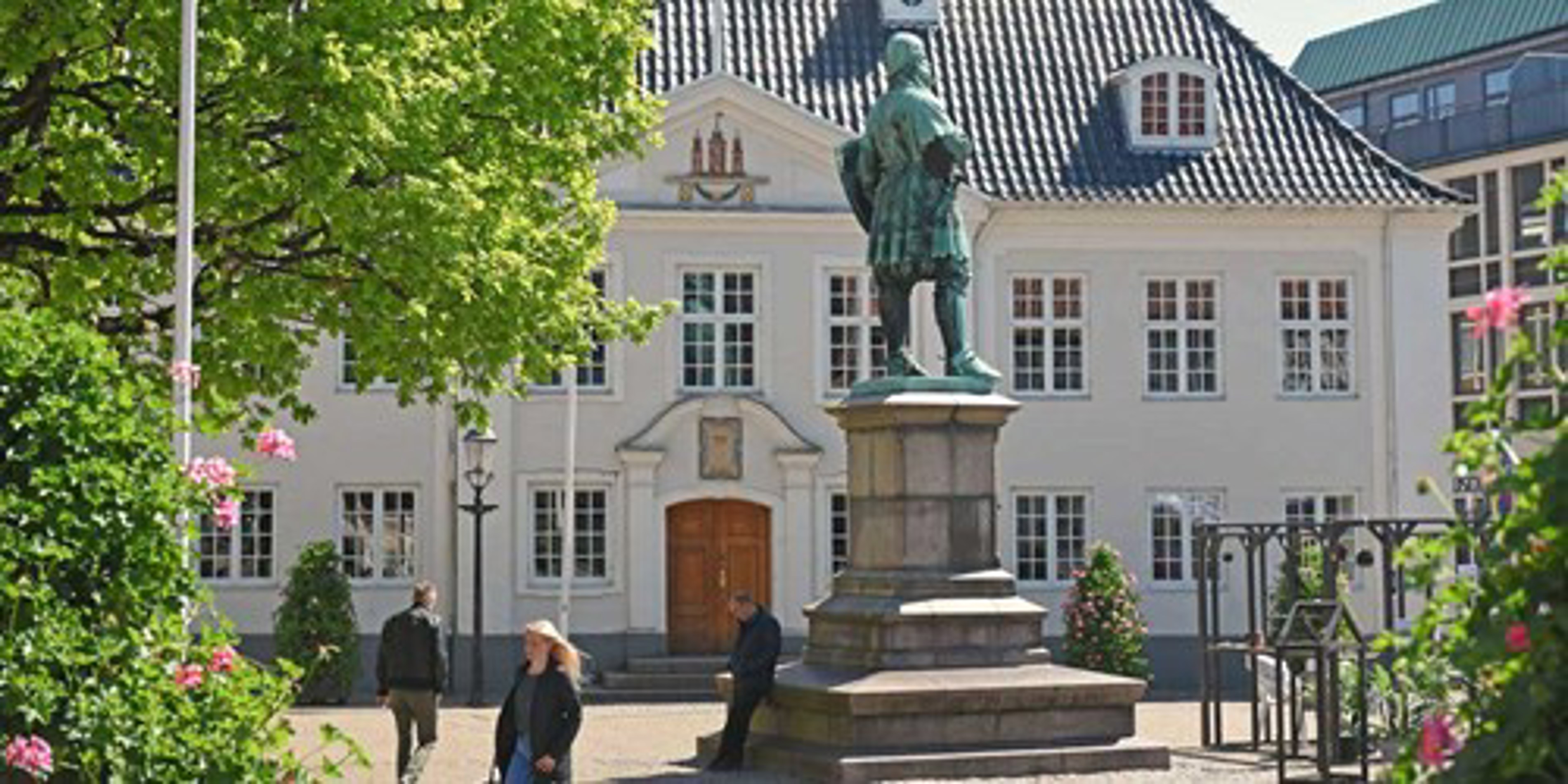 Statuen af mand foran historisk, hvid bygning med blåtag i bymiljø på solskinsdag.
