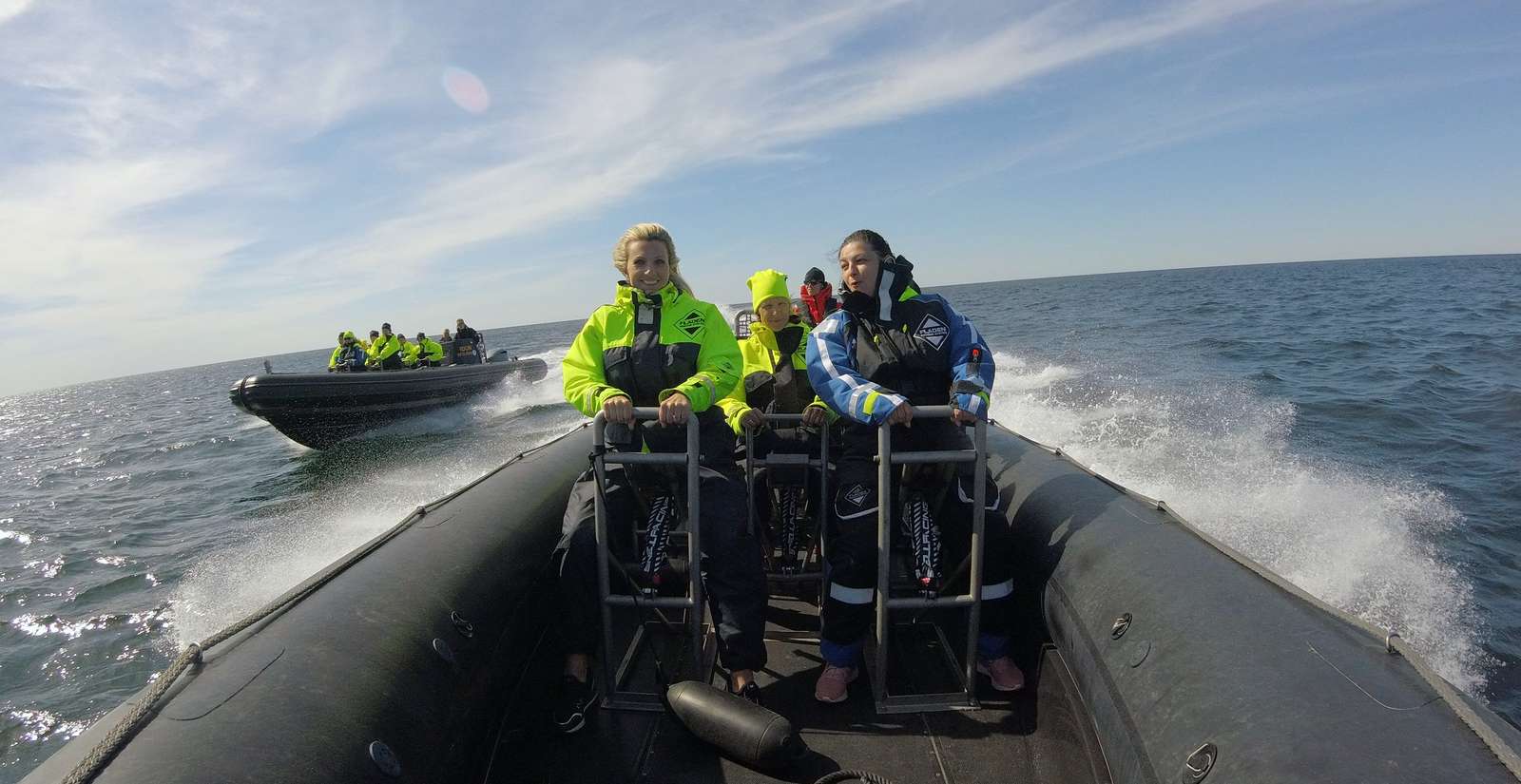 Personer iført lyse sikkerhedsjakker på en RIB-båd, der sejler hurtigt på åbent hav under en solrig himmel.