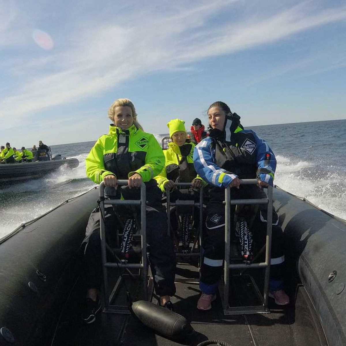 Personer iført lyse sikkerhedsjakker på en RIB-båd, der sejler hurtigt på åbent hav under en solrig himmel.