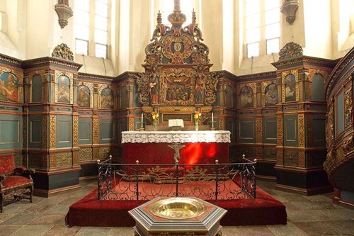 Dekoreret alter i en historisk kirke med træudskæringer og malerier, set fra kirkeskibets perspektiv.