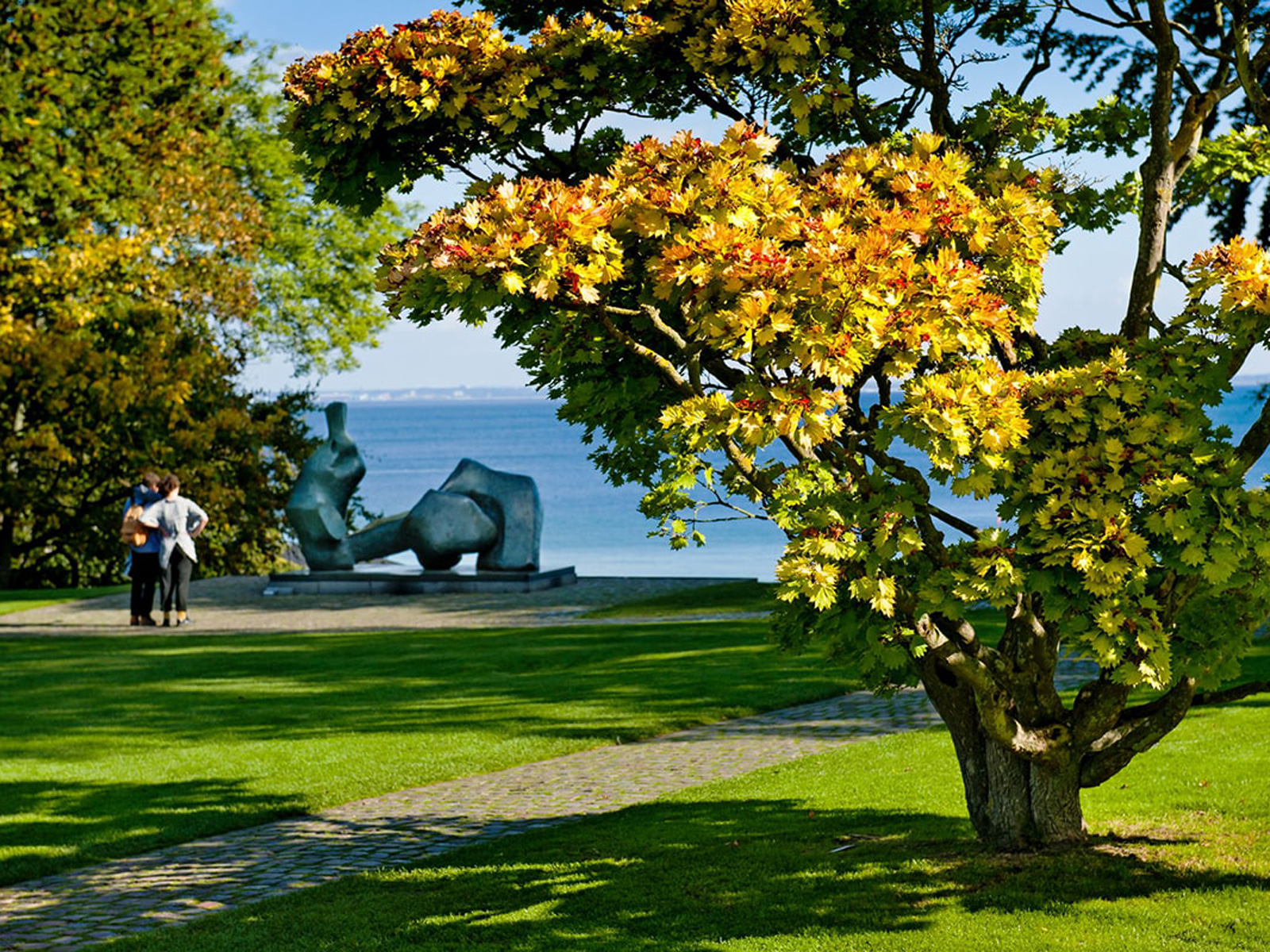 Skulptur i parken på Louisiana konstmuseum med utsikt mot havet och grönskande träd i förgrunden.