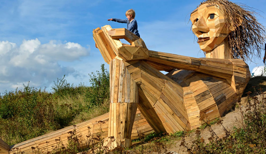 Jättestaty i trä, Kph Jättetrollen Trine, med ett barn som klättrar på statyns knä i en grönskande utomhusmiljö.