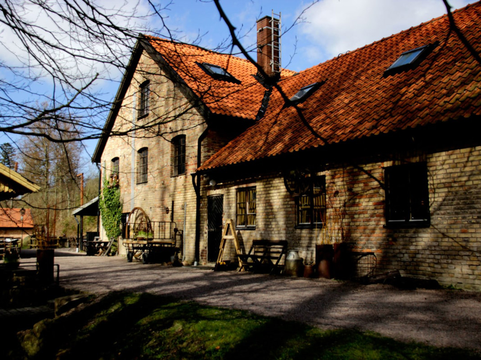 Traditionel murstensbygning med rødt tegltag belyst af sollys omgivet af træer og grusvej i en landlig idyl.