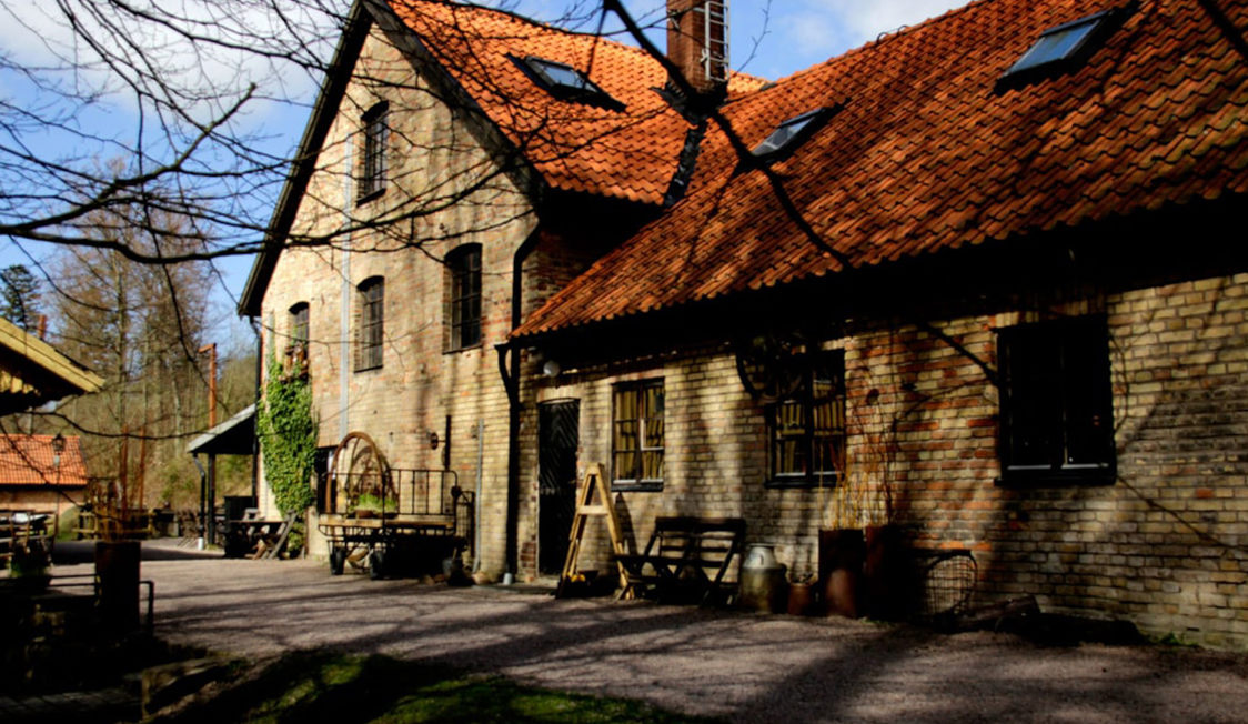 Traditionel murstensbygning med rødt tegltag belyst af sollys omgivet af træer og grusvej i en landlig idyl.