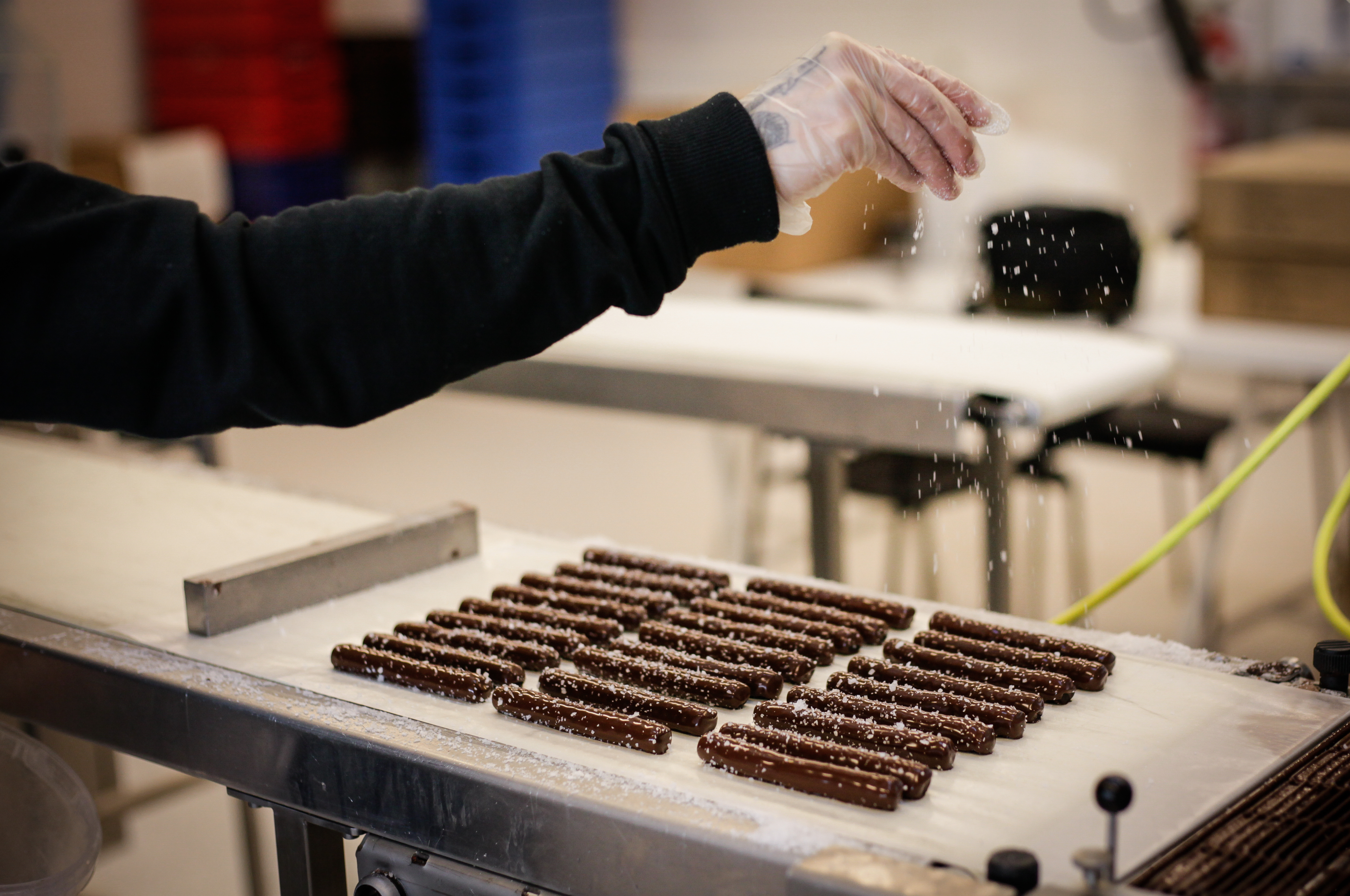 Hånd drysser sukker over chokoladebarer på bånd i produktionsmiljø.