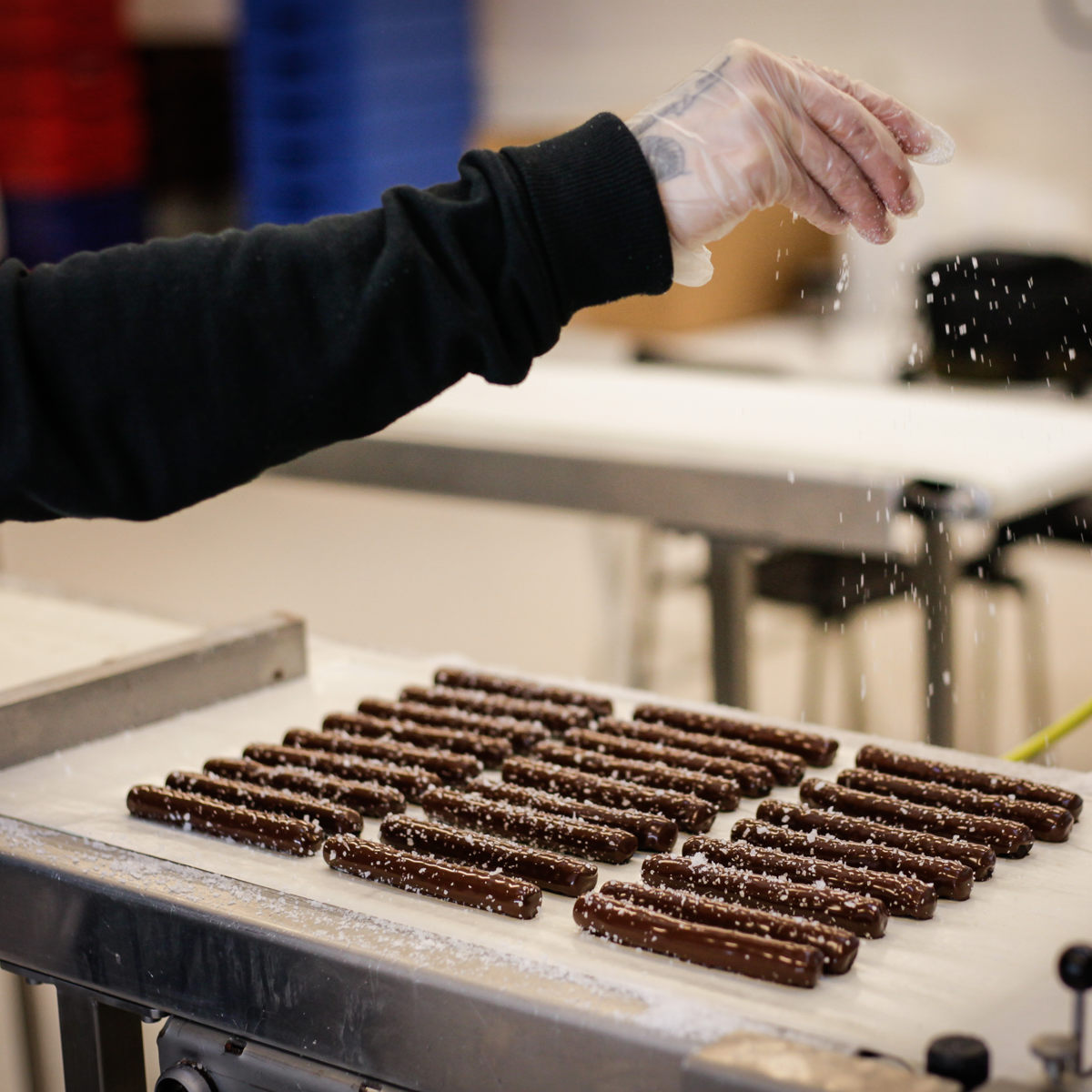 Hånd drysser sukker over chokoladebarer på bånd i produktionsmiljø.