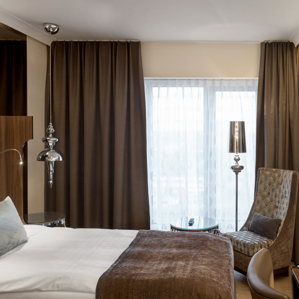Elegant hotelværelse med brun sengeløber, polstret stol, sølvfarvet lampe og store vinduer med gardiner.