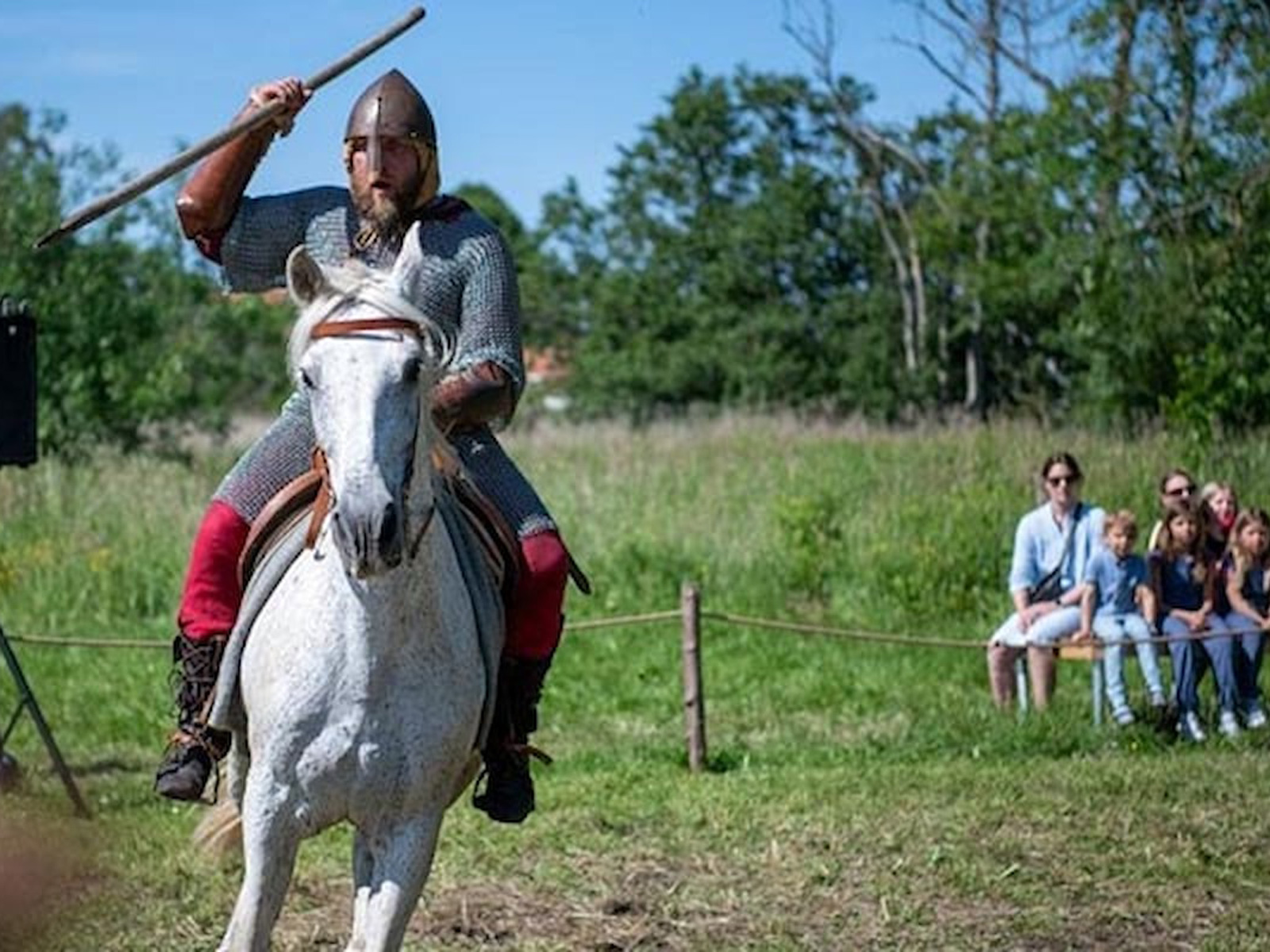 Riddare på vit häst i rustning under en Esrum Riddare-show, framför en publik på en somrig gräsäng.