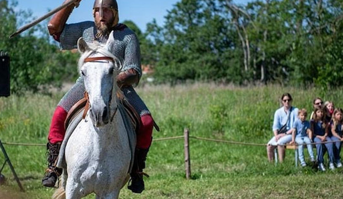 Riddare på vit häst i rustning under en Esrum Riddare-show, framför en publik på en somrig gräsäng.