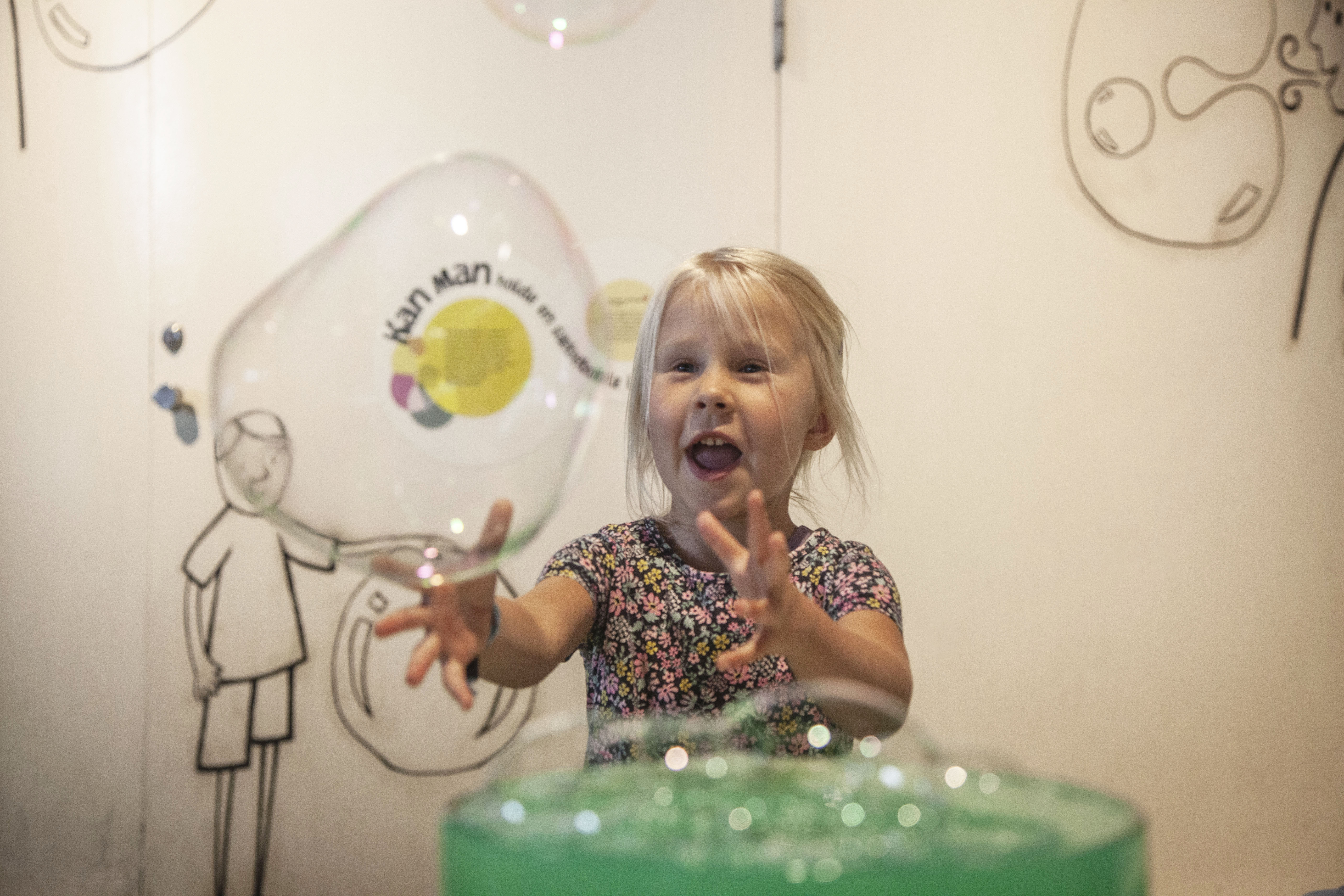 Litet barn leker glatt med en stor såpbubbla inomhus, omgiven av illustrerade väggmålningar.
