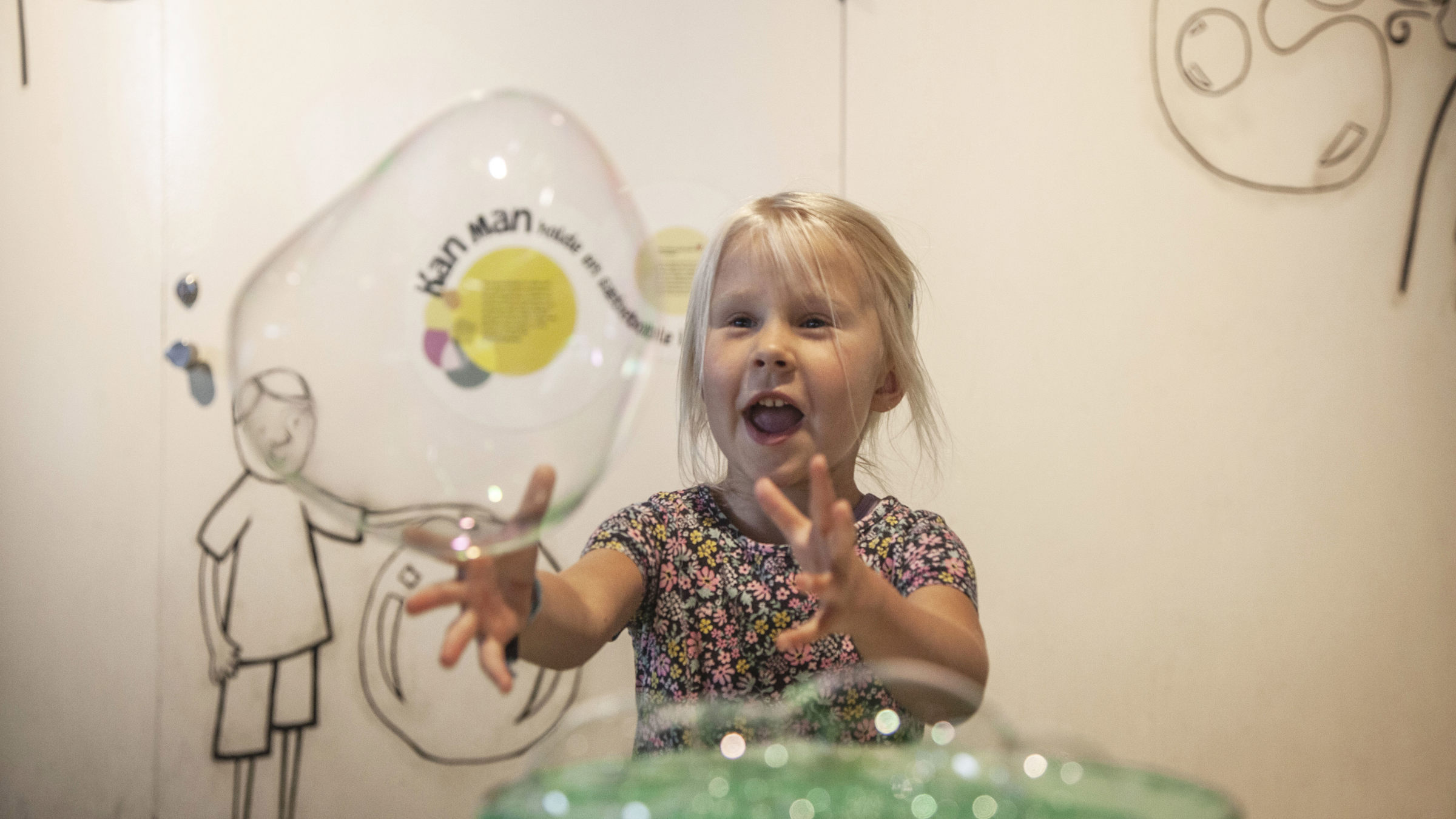 Litet barn leker glatt med en stor såpbubbla inomhus, omgiven av illustrerade väggmålningar.