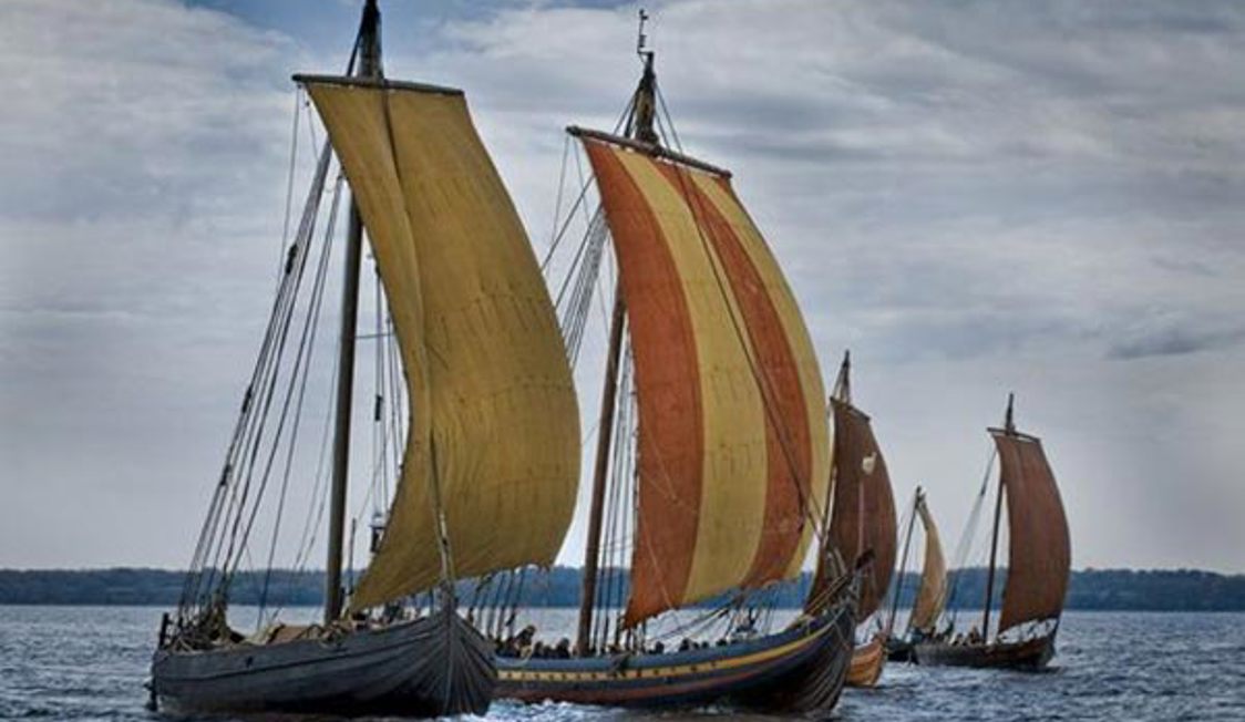 Vikingeskibe med farverige sejl seglar på roligt hav under molnig himmel, sedd från sidan.