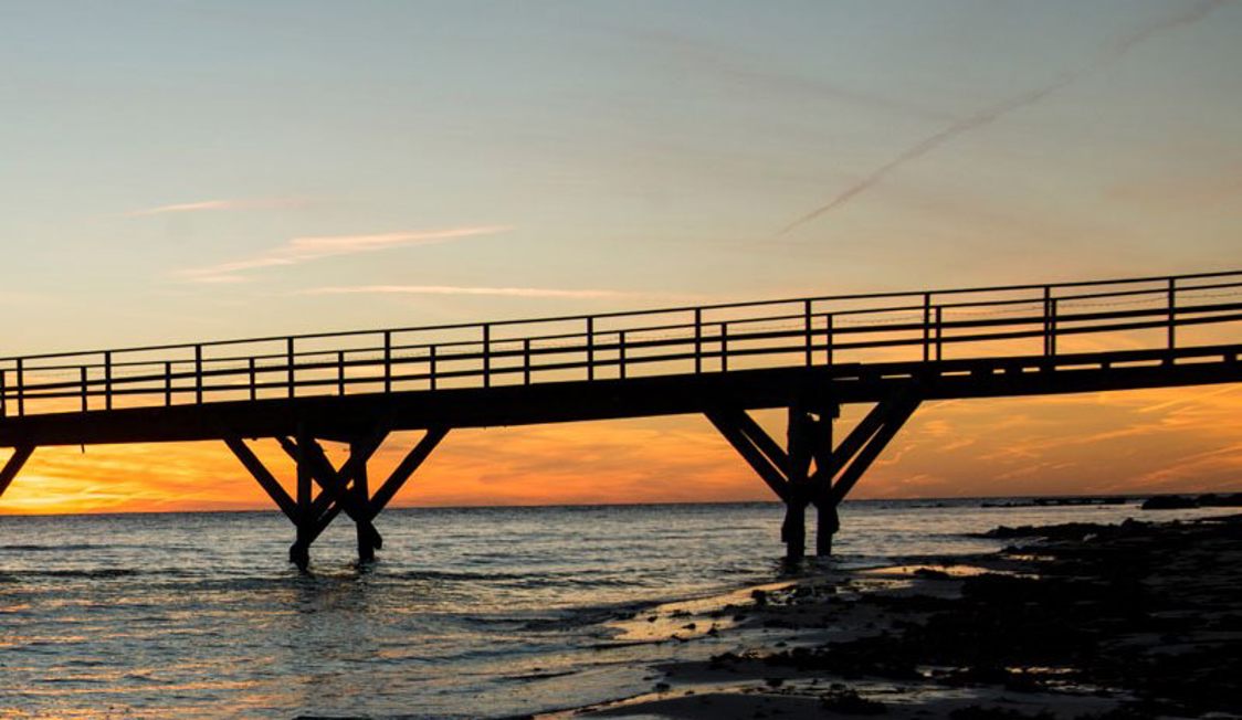 Træbro strækker sig over vandet i solnedgangen med orange og pink nuancer på himlen.