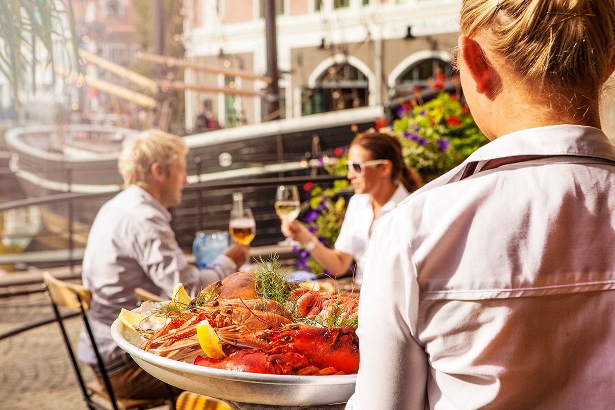 To personer sidder ved et cafébord i solen og drikker vin, mens en trjener er på vej over til dem med skaldyr.