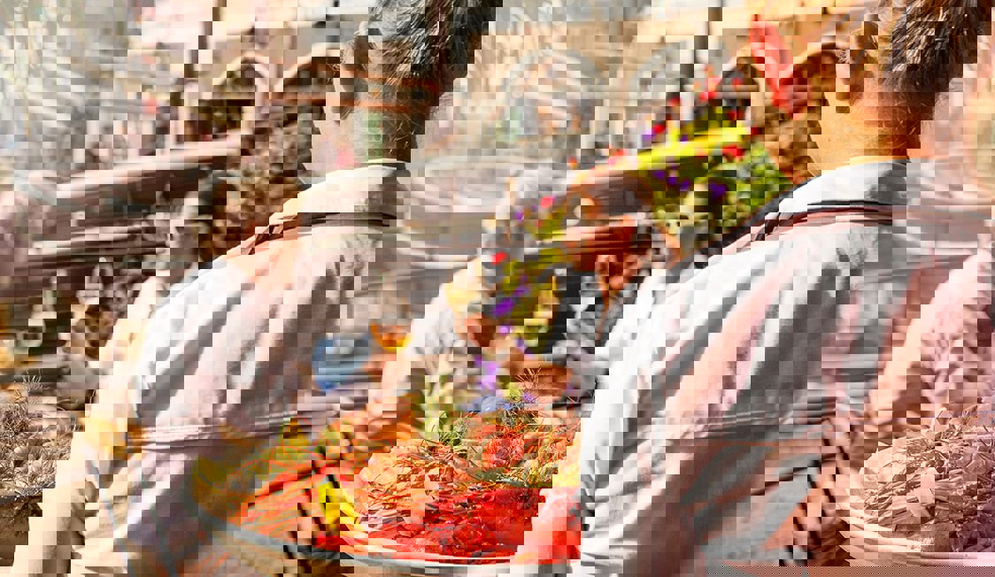 To personer sidder ved et cafébord i solen og drikker vin, mens en trjener er på vej over til dem med skaldyr.