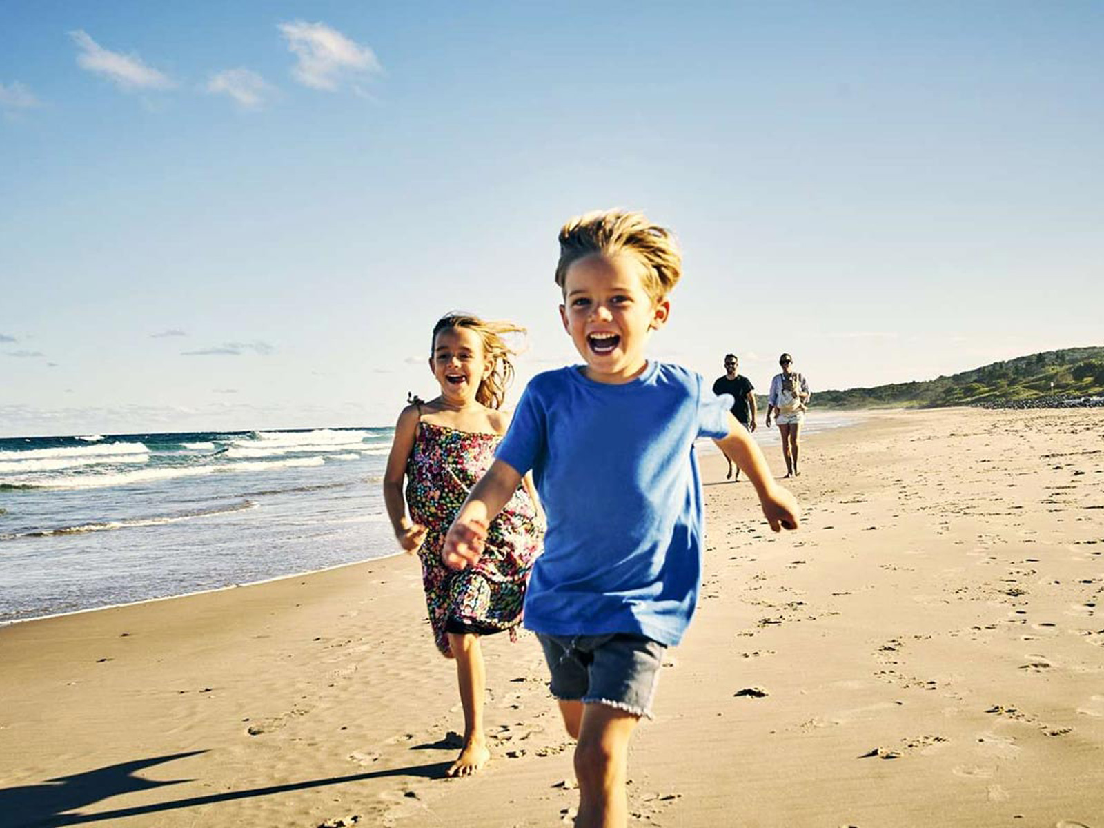 Barn springer glatt på stranden i solskenet med havet i bakgrunden.