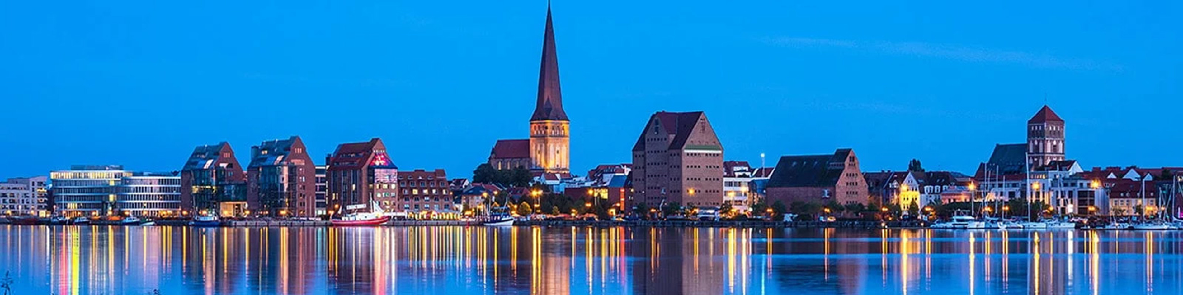 Rostocks nattliga skyline speglas i vattnet under en klar blå himmel.