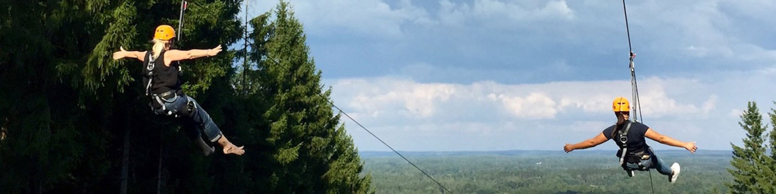 To personer svæver gennem skoven på en zipline i høj fart mens de bærer sikkerhedshjelme i et naturskønt område.