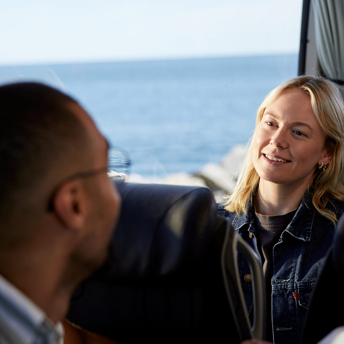 Kvinna och man samtalar på en buss med havsutsikt genom fönstret.